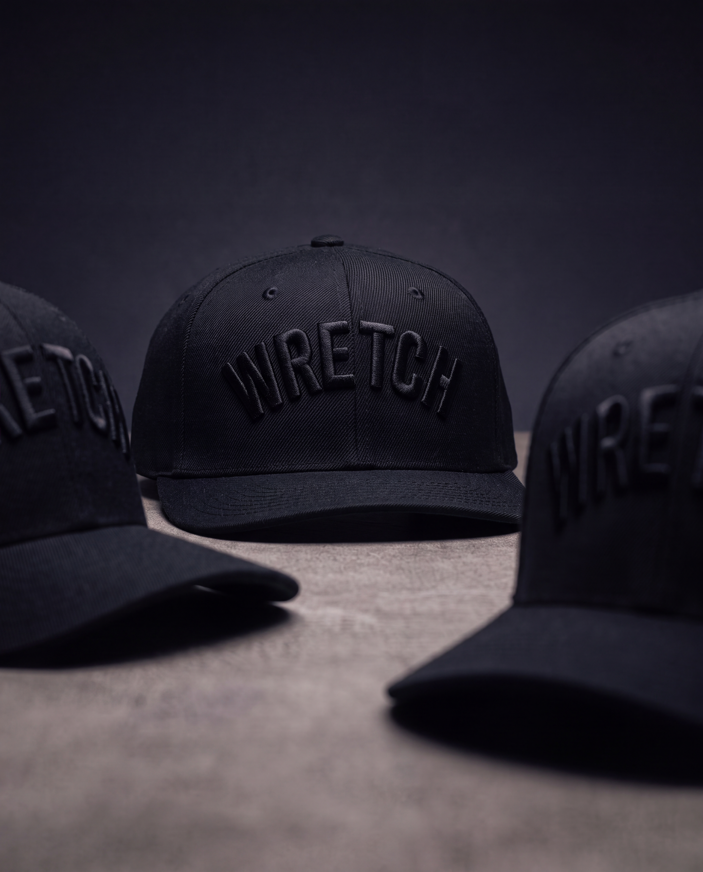 Three black baseball caps with embroidered 'WRETCH' branding, arranged on a textured surface against a dark studio background.