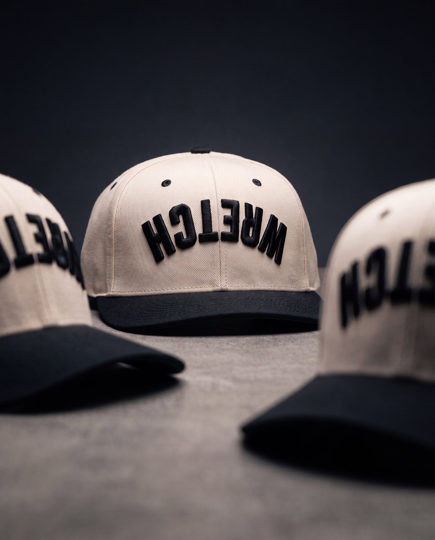 Three off-white snapback caps with black flat brims and black embroidered "WRETCH" on the front, arranged on a dark gray surface against a dark background.