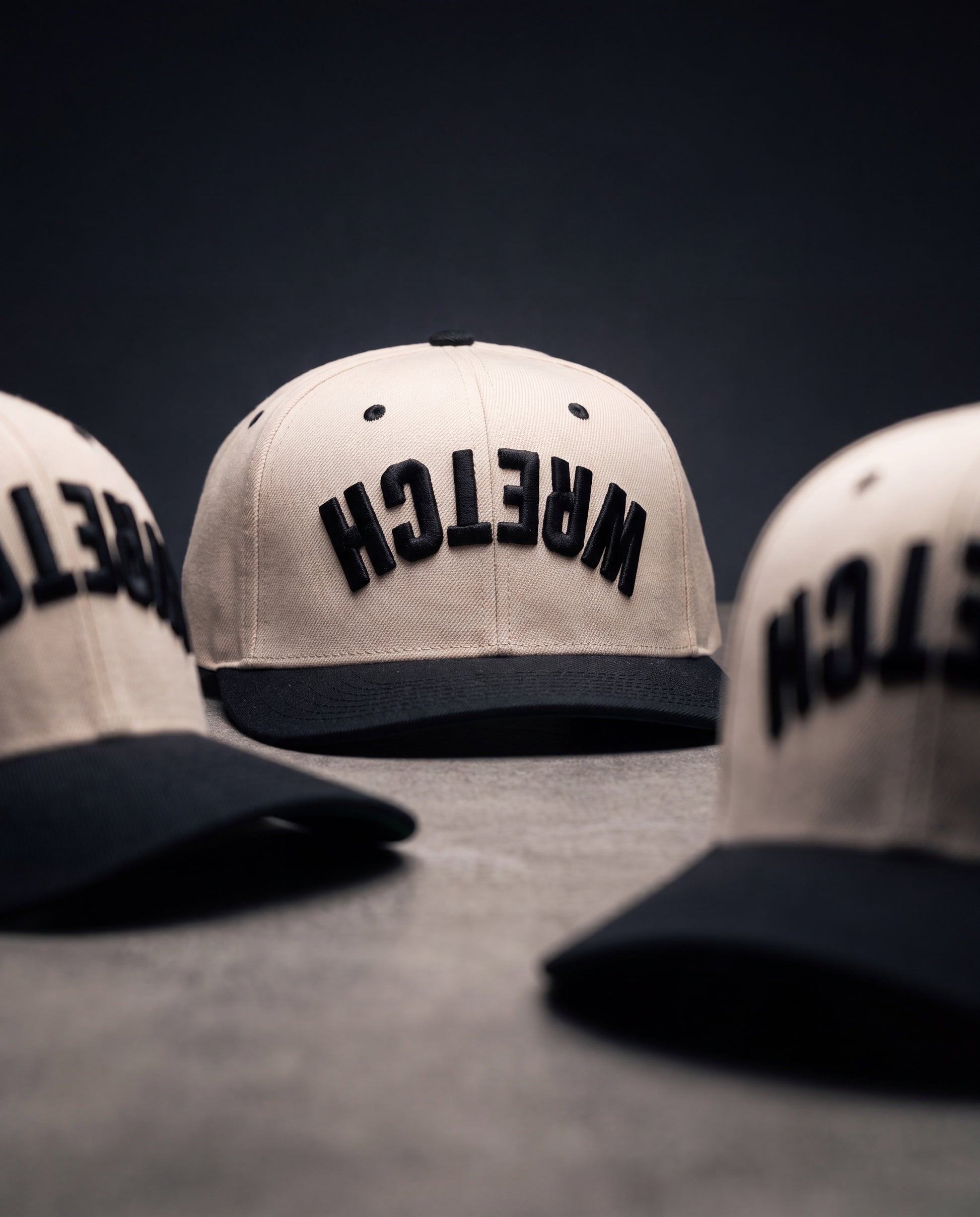 Three off-white snapback caps with black flat brims and black embroidered "WRETCH" on the front, arranged on a dark gray surface against a dark background.